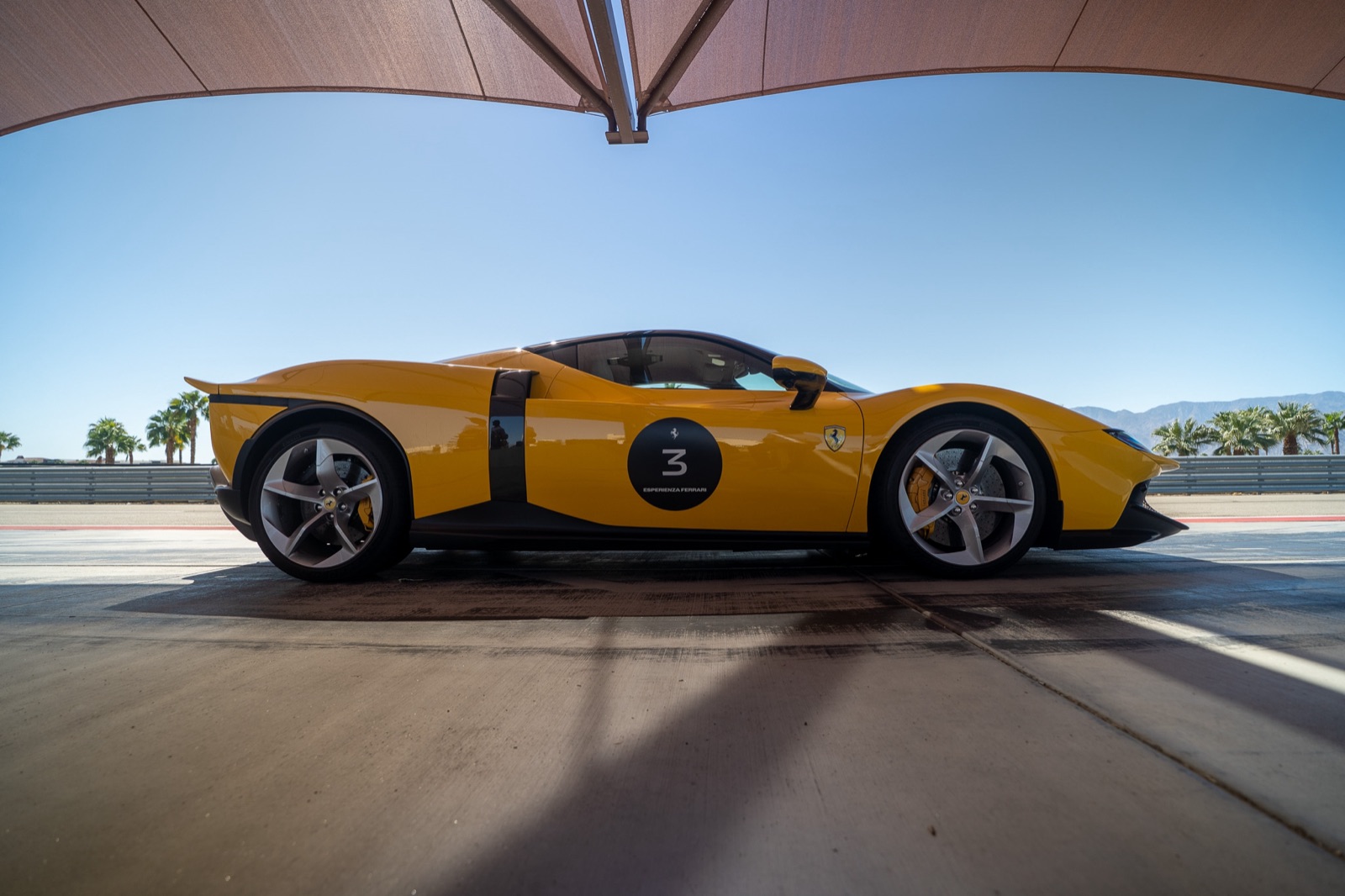 Side profile of a yellow Ferrari under a shade canopy in the pit lane, with palm trees and mountains beyond