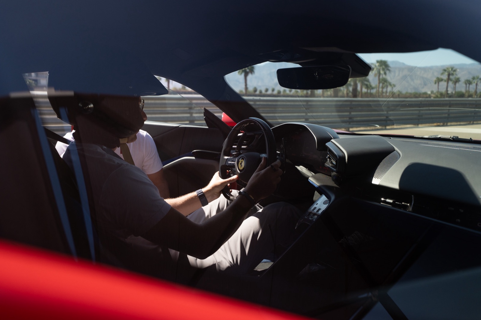 View through a Ferrari's side window of a helmeted driver gripping the steering wheel, the track wall and palm trees outside